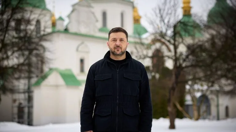 Wołodymyr Zełenski w czarnej kurtce stoi zimą na śniegu przed cerkwią ze złotymi kopułami w Kijowie