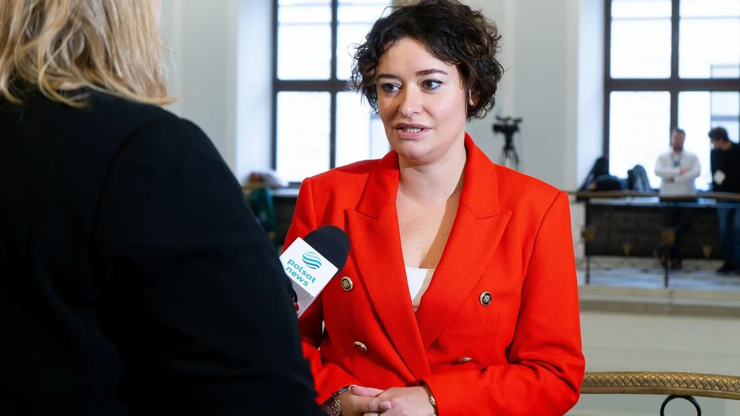 Anna Maria Żukowska w czerwonej marynarce udziela wywiadu dziennikarzom, mówiąc do mikrofonu.
