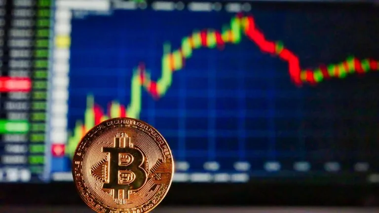 Moneta Bitcoin na pierwszym planie, za nią ekran z wykresem notowań, na którym widać spadki i wzrosty kursu.
