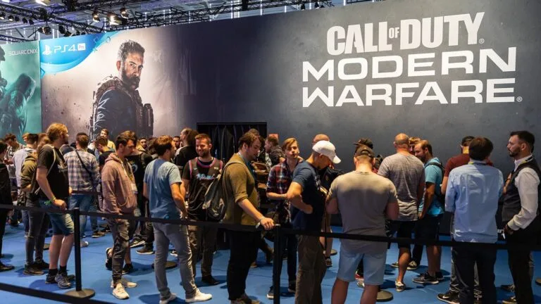 Kolejka graczy przy stoisku Call of Duty: Modern Warfare na targach Gamescom, za nimi duży baner z logo gry i postacią.