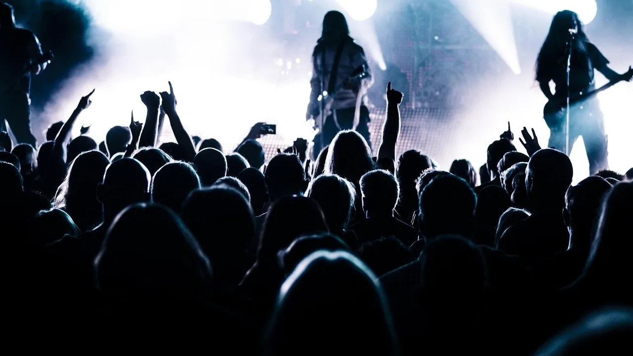 Tłum fanów pod sceną koncertu rockowego; na podświetlonej scenie widoczni muzycy z gitarami.