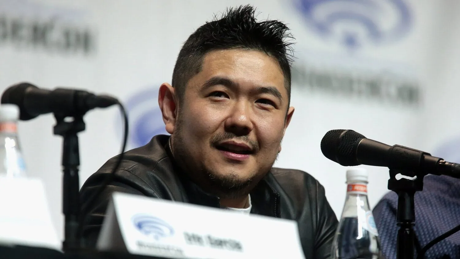 Eric Bauza w czarnej skórzanej kurtce siedzi przy stole panelowym i mówi do mikrofonu na WonderCon w Anaheim w 2018 roku.