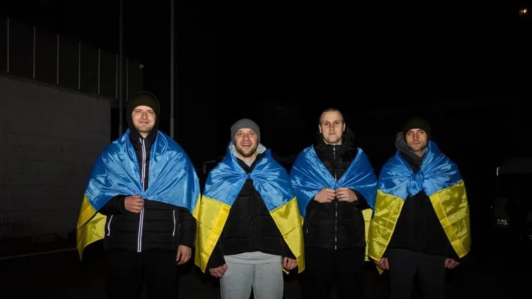 Czterech mężczyzn owiniętych flagami Ukrainy stoi nocą na zewnątrz po wymianie jeńców z Rosją