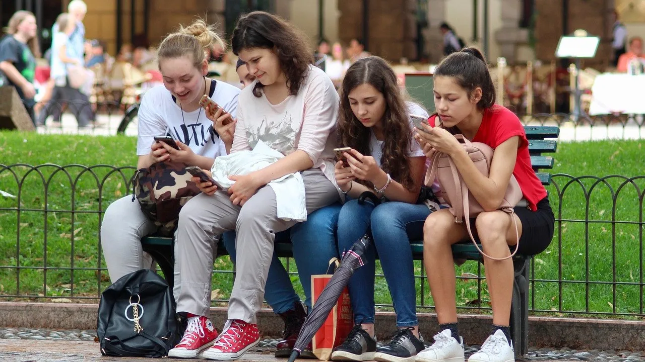 Cztery nastolatki siedzą na ławce w parku i przeglądają smartfony.