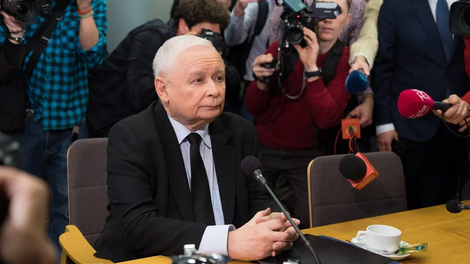 Jarosław Kaczyński siedzi przy stole z mikrofonami podczas konferencji prasowej, za nim dziennikarze z kamerami.