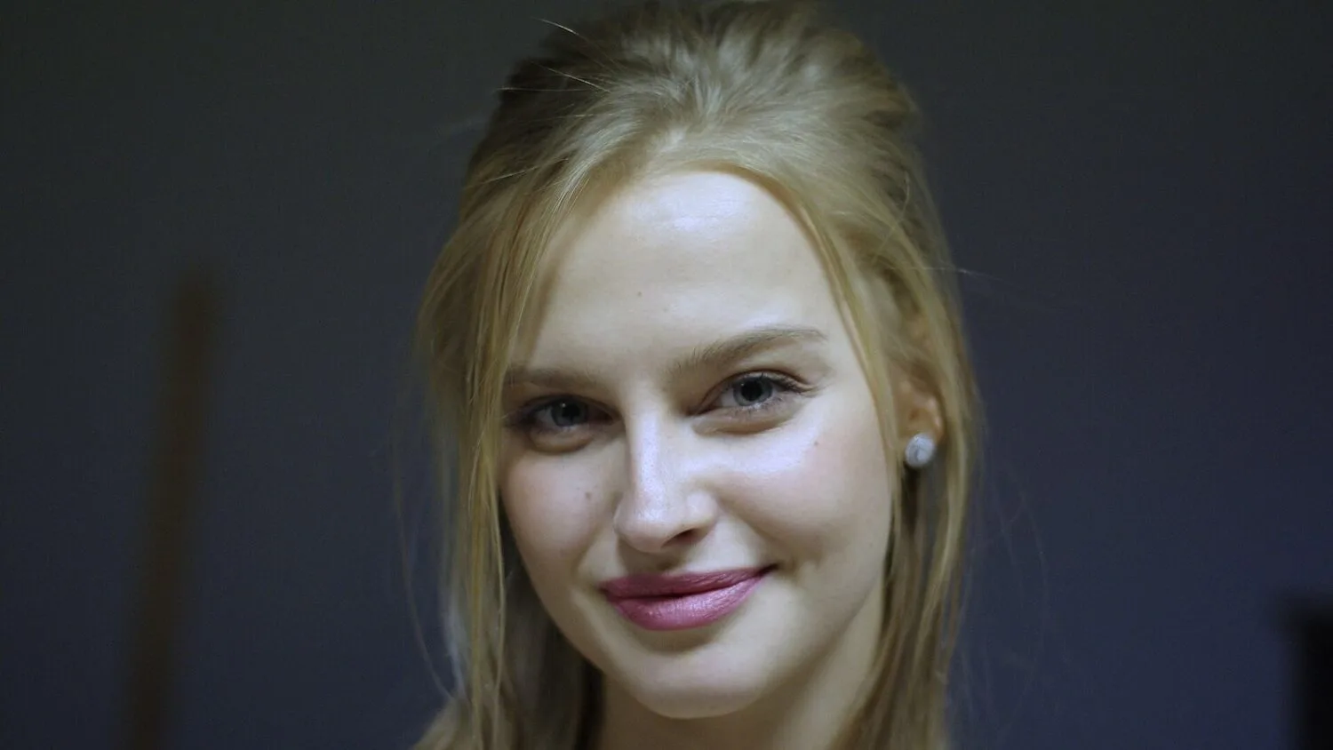 Karolina Pisarek, blondynka z rozpuszczonymi włosami i perłowym kolczykiem, uśmiecha się do kamery na ciemnym tle.