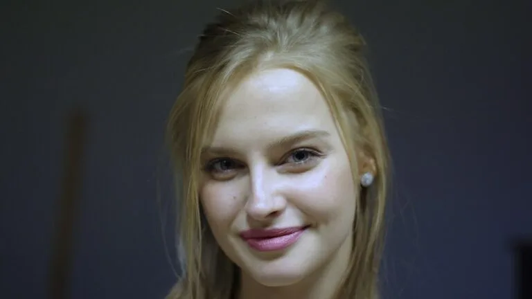 Karolina Pisarek, blondynka z rozpuszczonymi włosami i perłowym kolczykiem, uśmiecha się do kamery na ciemnym tle.