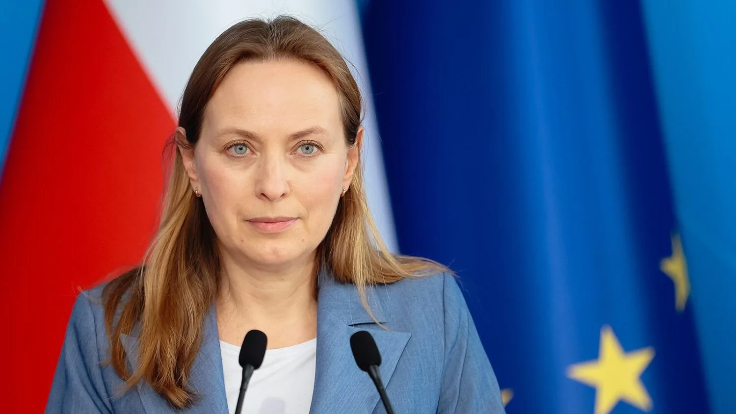 Katarzyna Pełczyńska-Nałęcz przy mikrofonach na konferencji prasowej, na tle flag Polski i UE.