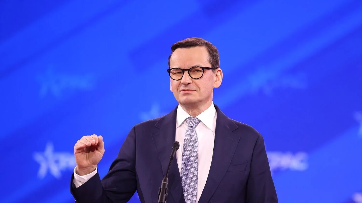 Mateusz Morawiecki w ciemnym garniturze i okularach przemawia za szklanym pulpitem na niebieskim tle konferencji CPAC