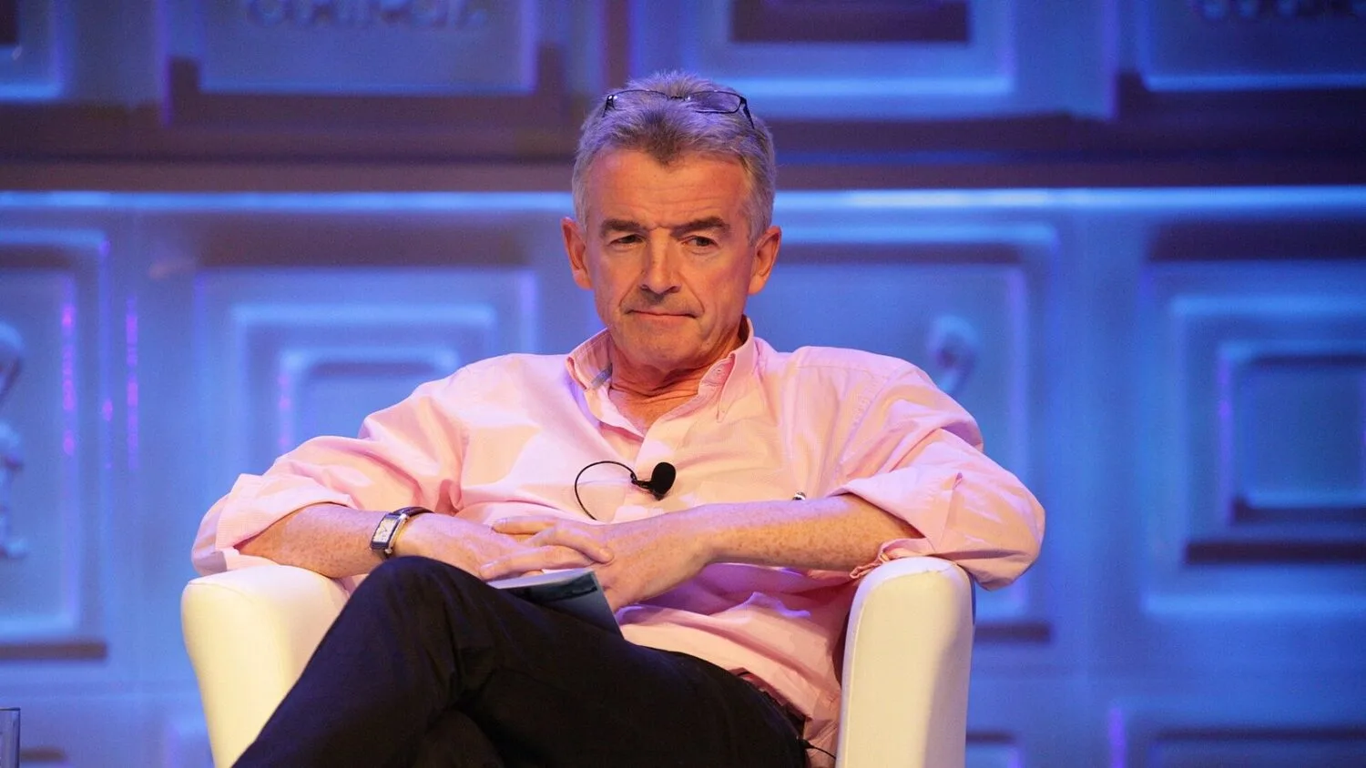 Michael O'Leary, szef Ryanaira, siedzi w białym fotelu na scenie konferencji w różowej koszuli.