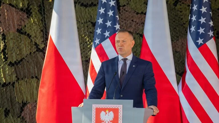 Karol Nawrocki przemawia przy mównicy z godłem Polski, na tle flag Polski i USA, podczas uroczystości w Oleszni.