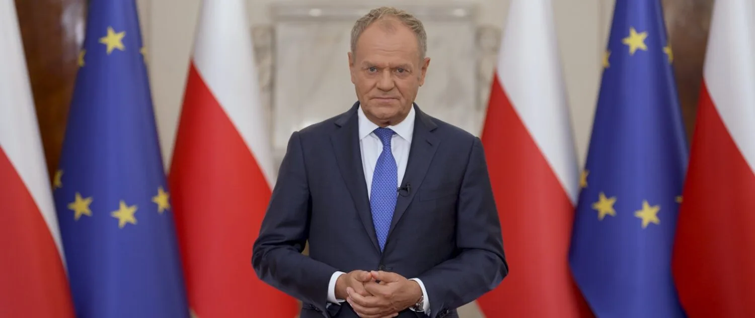 Premier Donald Tusk stoi przed flagami Polski i Unii Europejskiej, z dłońmi splecionymi przed sobą, podczas nagrania orędzia.