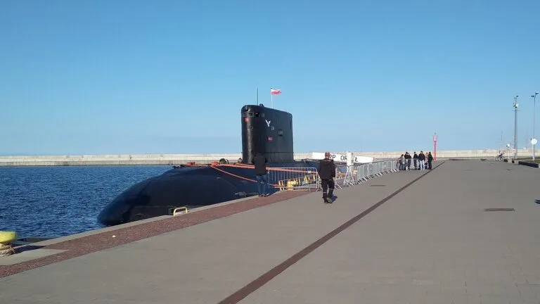 Polski okręt podwodny ORP Orzeł przy nabrzeżu Skweru Kościuszki w Gdyni; na promenadzie przechodnie, w tle barierki i morze.
