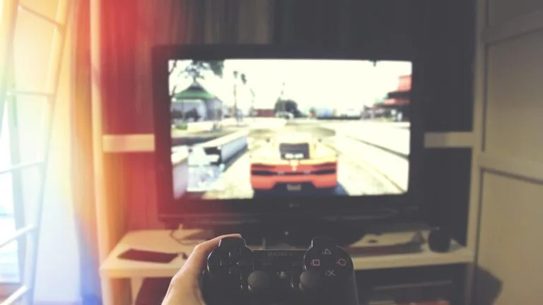 Pad PlayStation w dłoni przed telewizorem, na ekranie gra samochodowa w mieście
