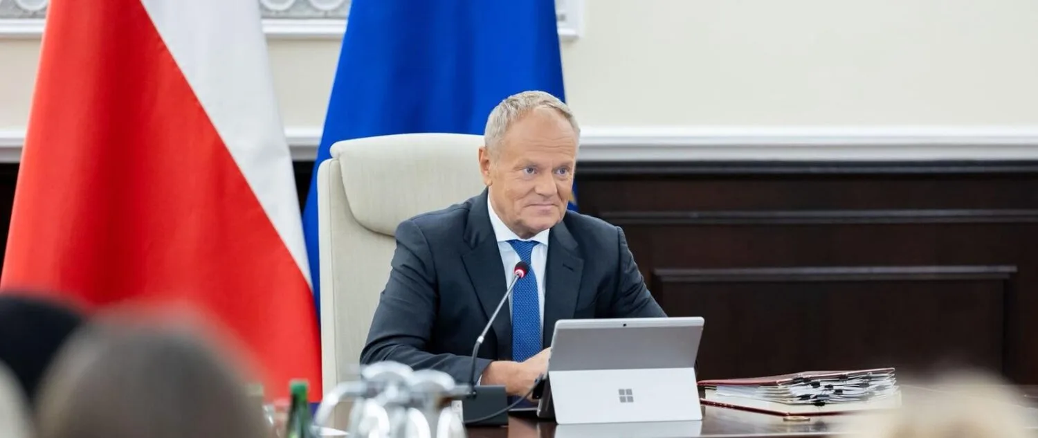 Premier Donald Tusk podczas posiedzenia rządu przy stole, z laptopem i mikrofonem; w tle flagi Polski i Unii Europejskiej