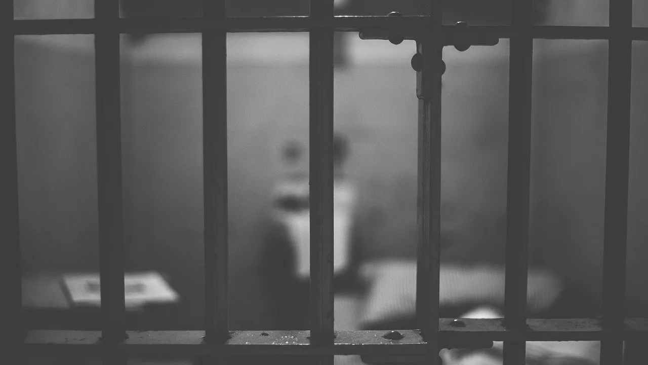 Czarno-biała fotografia: metalowe kraty celi więziennej, w tle niewyraźne wnętrze z łóżkiem.