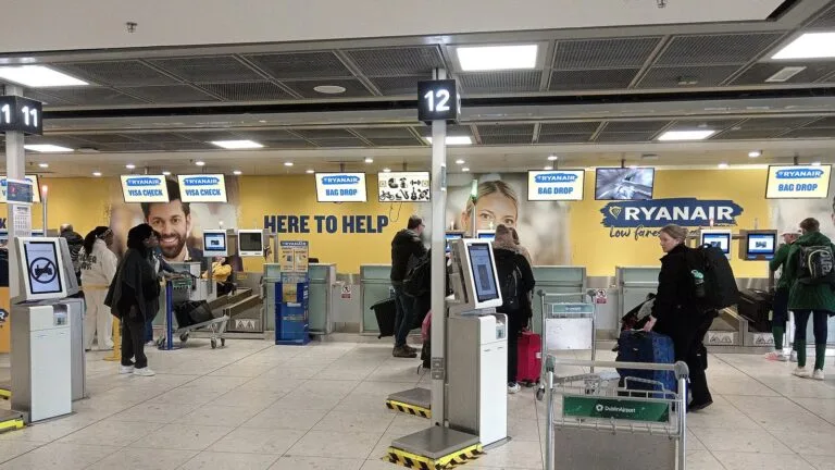 Stanowiska odprawy bagażu Ryanaira na lotnisku w Dublinie; pasażerowie z walizkami czekają w kolejce do nadania bagażu.