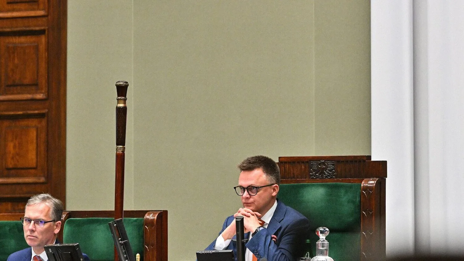Szymon Hołownia siedzi w prezydium Sejmu podczas obrad, z ręką przy brodzie, w marynarce i okularach.