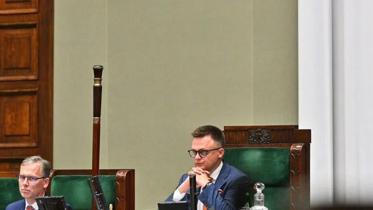Szymon Hołownia siedzi w prezydium Sejmu podczas obrad, z ręką przy brodzie, w marynarce i okularach.