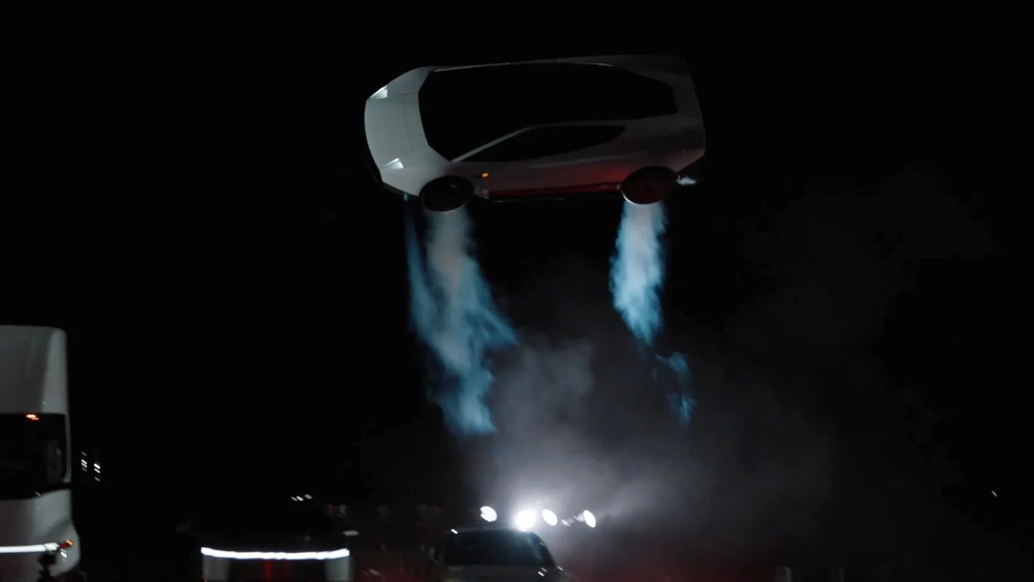 Biały Tesla Roadster unosi się w powietrzu nad parkingiem, pod nim dwa strumienie gazu z dysz, obok ciężarówka Tesla Semi