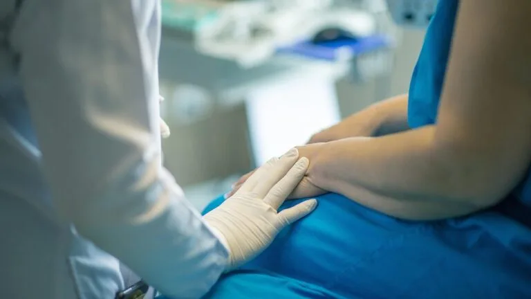 Lekarz w białym fartuchu i rękawiczkach trzyma dłoń pacjenta w niebieskim stroju, leżącego na łóżku w szpitalu.