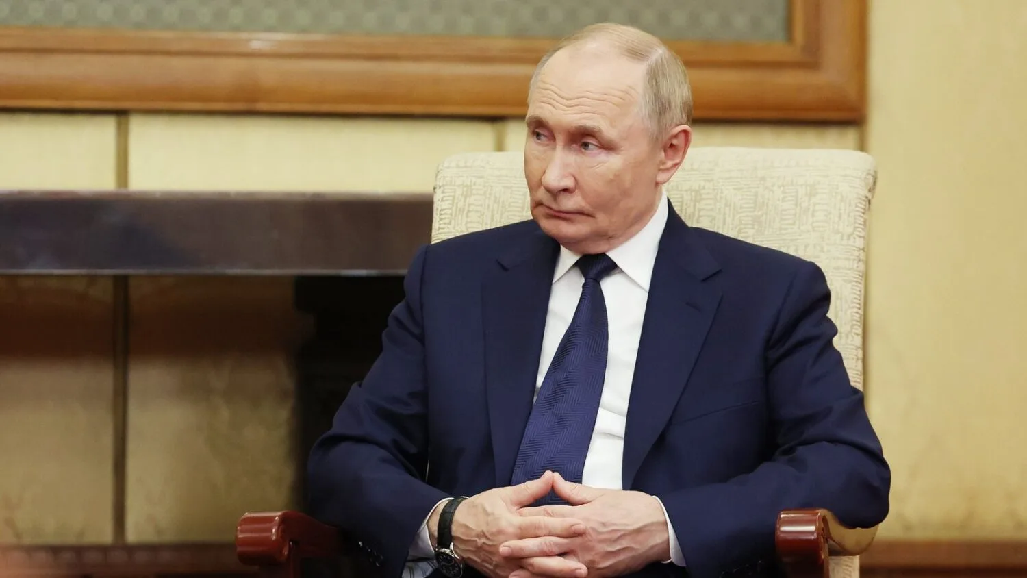 Władimir Putin siedzi w fotelu, w ciemnym garniturze i krawacie, ze splecionymi dłońmi i poważnym wyrazem twarzy