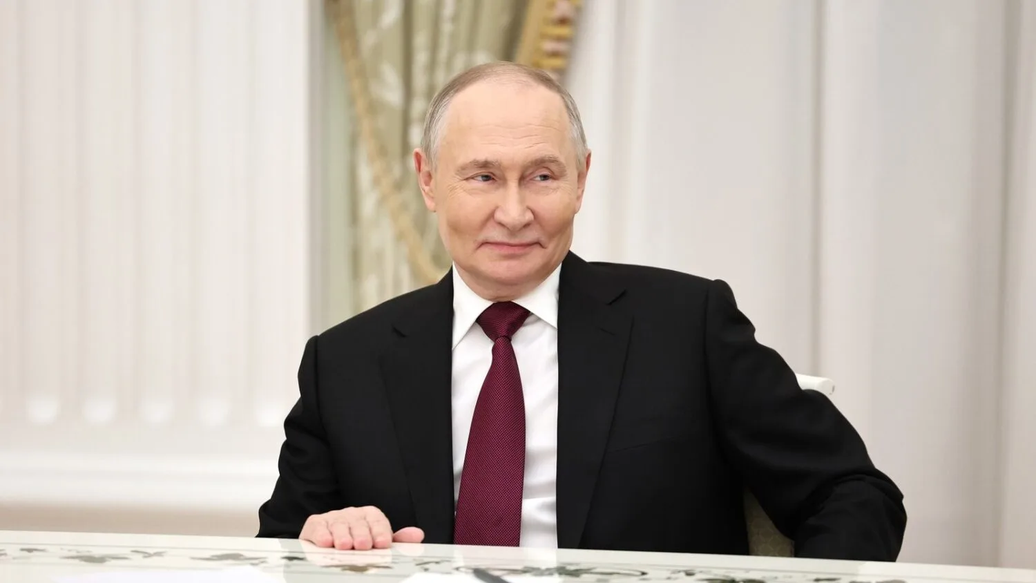 Władimir Putin siedzi przy stole w ciemnym garniturze i czerwonym krawacie, w jasnej sali na Kremlu.